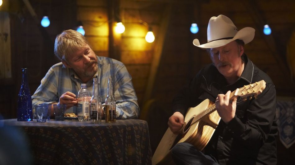 Två män vid ett bord, en av dem spelar gitarr. Ur filmen Det enda rationella.